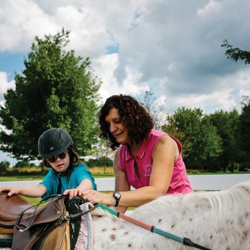 An equine assisted services volunteer helps a student mount a spotted pony